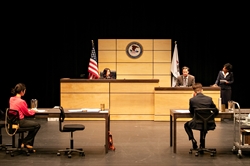 [ai] A courtroom scene with two attorneys seated at desks, a judge presiding at the front, and a witness on the stand. The American flag is displayed. The setting has a modern design with wooden panels.